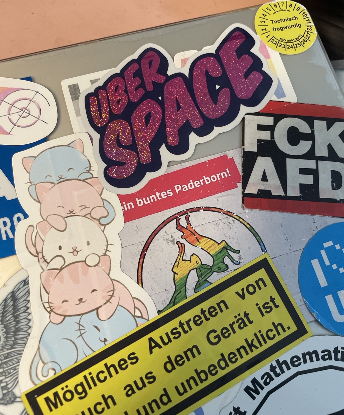 Purple shiny Uberspace sticker on a laptop, around it some stickers in german language: FCK AFD, Technically questionable, 5 cats lying on top of each other in the colors of the trans flag, covered sticker on which it says …colorful Paderborn, “Possible leakage of smoke from the device is normal and harmless”